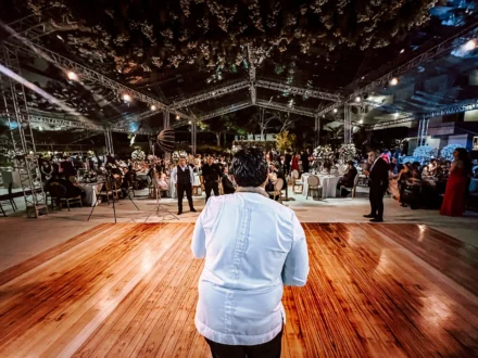 Vista desde atrás de un músico vestido de blanco sobre un escenario de madera, frente a un público en un evento nocturno elegante con mesas decoradas y luces bajo una estructura cubierta transparente.
