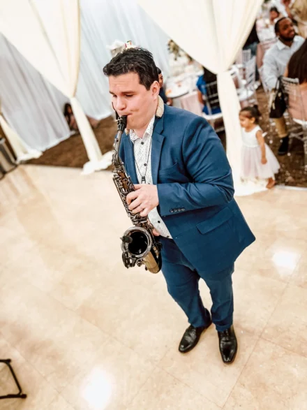 Iván De León, saxofonista profesional, ambientando una boda elegante con su música. Vestido con un traje azul y camisa blanca de detalles, interpreta una melodía especial mientras los invitados disfrutan del evento en un salón decorado con tonos blancos y dorados.