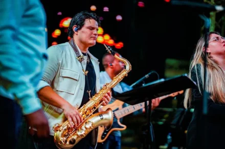 Iván De León, saxofonista profesional, tocando su saxofón dorado durante un concierto en vivo. Con una expresión concentrada y auriculares in-ear, se encuentra acompañado de una banda, mientras las luces del escenario crean un ambiente vibrante y enérgico. Ideal para representar música en vivo y presentaciones de jazz o fusión.