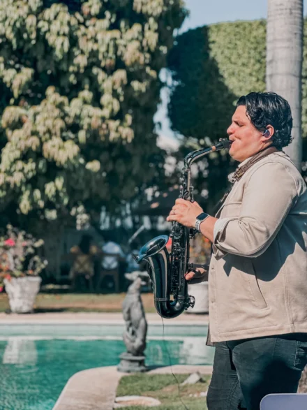 Iván De León, saxofonista profesional, tocando su saxofón negro en un evento al aire libre junto a una piscina. Viste una chaqueta beige con pantalón oscuro y utiliza auriculares in-ear mientras interpreta su música con el fondo de un jardín exuberante. Una escena que refleja elegancia y armonía con la naturaleza.