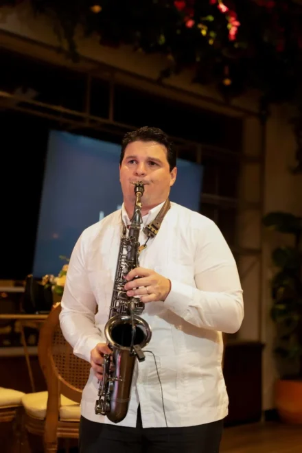 Iván De León, saxofonista profesional, interpretando su música en un evento cultural. Con una guayabera blanca y un saxofón negro, transmite elegancia y tradición mientras actúa en un escenario con una iluminación cálida y detalles decorativos naturales. Una imagen que resalta su talento en presentaciones en vivo.