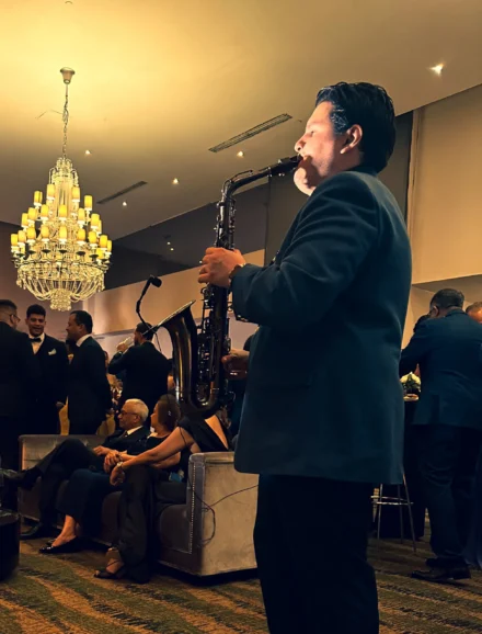 Iván De León, saxofonista profesional, interpretando su música en un evento exclusivo de gala. Con un elegante traje oscuro, toca su saxofón negro en un salón lujosamente decorado con un imponente candelabro y un público distinguido disfrutando del ambiente sofisticado. La imagen transmite clase y elegancia musical.
