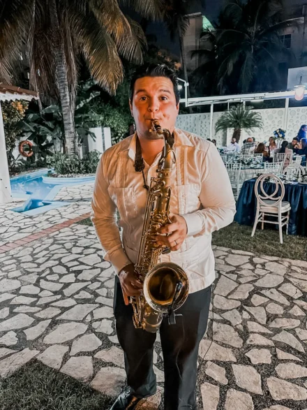 Iván De León, saxofonista profesional, tocando su saxofón dorado en un evento nocturno al aire libre. Vestido con una guayabera blanca y pantalón oscuro, su interpretación crea un ambiente especial en una fiesta junto a una piscina, rodeado de palmeras y luces decorativas. La música en vivo eleva la experiencia del evento.