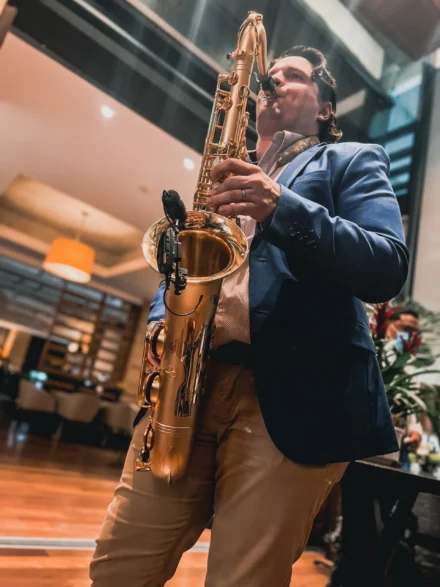 Iván De León, talentoso saxofonista, interpretando su música en una presentación exclusiva en un elegante restaurante. Con un traje azul y pantalón beige, su saxofón dorado brilla bajo la iluminación cálida del lugar, creando un ambiente sofisticado y envolvente para los asistentes.