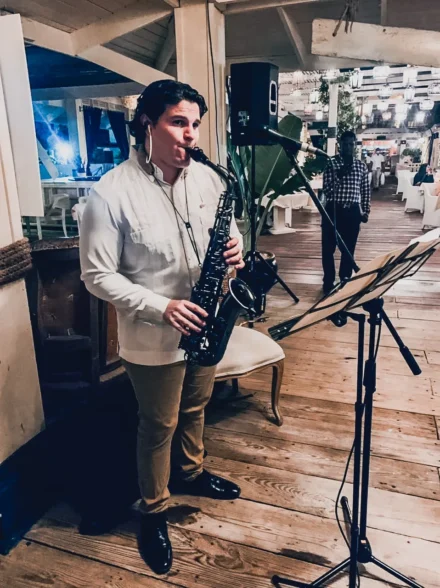 Iván De León, saxofonista profesional, tocando su saxofón negro en un elegante restaurante. Viste una guayabera blanca y pantalón beige, mientras interpreta su música con partituras en un atril. El ambiente cálido con iluminación tenue y decoración rústica en madera crea una atmósfera íntima y sofisticada.