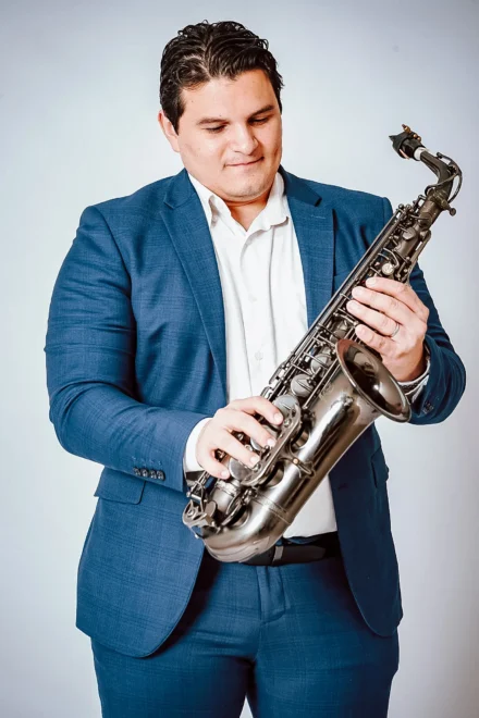 Iván De León, saxofonista profesional, en una sesión de fotos de estudio con fondo blanco. Viste un elegante traje azul y camisa blanca mientras sostiene su saxofón negro con una expresión de apreciación y concentración. La imagen transmite profesionalismo y pasión por la música.