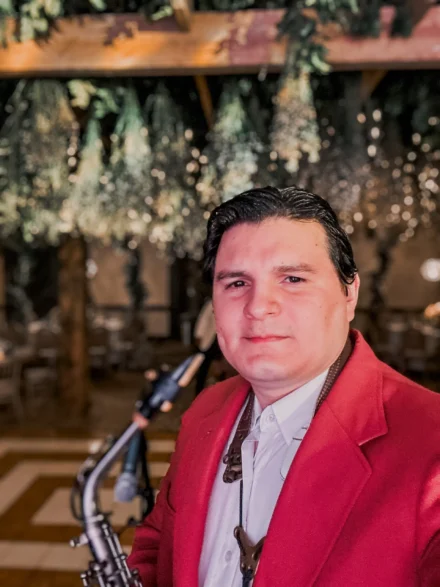 Iván De León, saxofonista profesional, en un evento de gala con un elegante traje rojo y camisa blanca. Con un saxofón en primer plano y un fondo decorado con luces y follaje colgante, la imagen transmite un ambiente sofisticado y festivo, resaltando su presencia musical en eventos exclusivos.