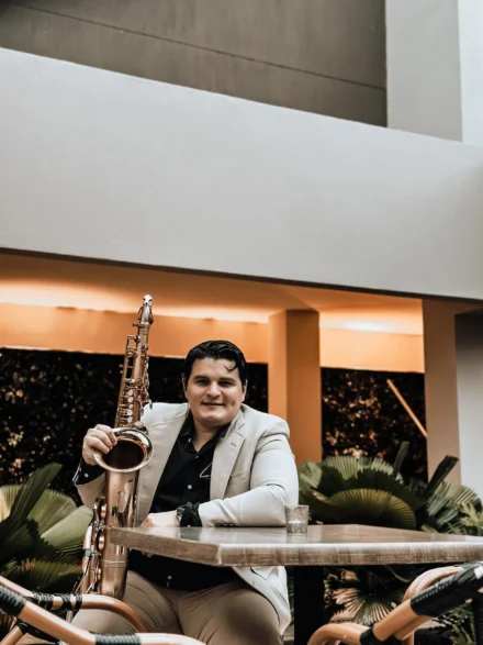 Iván De León, saxofonista profesional, en un ambiente relajado, sentado en una terraza con un saxofón dorado apoyado en la mesa. Viste un traje beige con camisa negra, transmitiendo elegancia y confianza. La iluminación cálida y el entorno moderno crean una escena sofisticada y acogedora