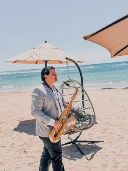 Iván De León, saxofonista profesional, caminando por la arena con su saxofón dorado en una presentación diurna en la playa. Viste un traje gris claro con camisa blanca, mientras el sol ilumina el mar azul y las sombrillas crean un ambiente relajante y exclusivo. Una escena perfecta que combina música y naturaleza.