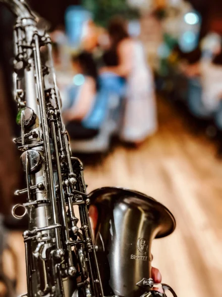 Primer plano de un saxofón negro brillante con detalles metálicos, destacando su estructura y marca ‘Brücker’. Al fondo, una escena borrosa de un evento elegante con invitados disfrutando del ambiente.